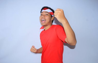 Heyecanlı Endonezyalı genç adamın portresi Endonezya 'daki başarısını ya da amacını kutlamak için kırmızı t-shirt ve saç bandı takarak yumruğunu kaldırdı. Genç adam başarısı ve memnuniyeti için evet işareti yapıyor.