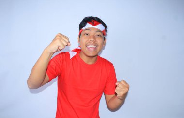 Heyecanlı Endonezyalı genç adamın portresi Endonezya 'daki başarısını ya da amacını kutlamak için kırmızı t-shirt ve saç bandı takarak yumruğunu kaldırdı. Genç adam başarısı ve memnuniyeti için evet işareti yapıyor.