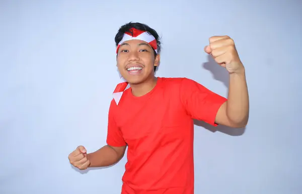 興奮したインドネシアの若い男性の肖像画は 赤いTシャツとヘッドバンドを着てインドネシア独立記念日の成功や目標を祝うために彼の拳を上げました 若い男は彼の達成と満足のためのイエスのジェスチャーを示します — ストック写真