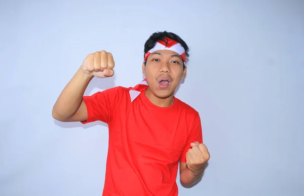 興奮したインドネシアの若い男性の肖像画は 赤いTシャツとヘッドバンドを着てインドネシア独立記念日の成功や目標を祝うために彼の拳を上げました 若い男は彼の達成と満足のためのイエスのジェスチャーを示します — ストック写真
