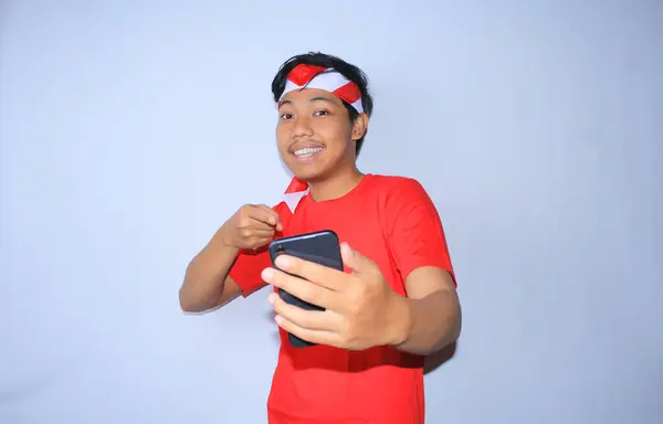 笑顔のインドネシアの若い男性は 白で隔離された赤いTシャツとヘッドバンドを身に着けている幸せで興奮した顔で自分の携帯電話を指摘 — ストック写真