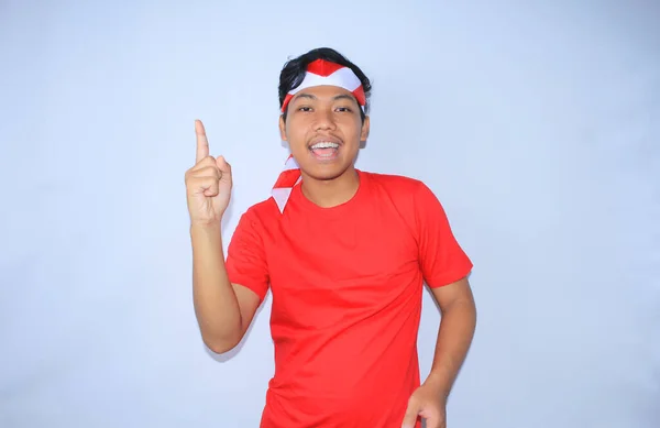 幸せなインドネシアの若い成人は笑顔で 白で隔離された赤いTシャツと国旗ヘッドバンドを身に着けている製品を提示するために指を指差しています — ストック写真
