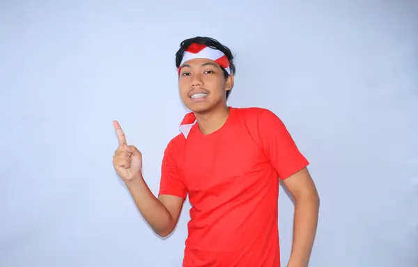 幸せなインドネシアの若い成人は笑顔で 赤いTシャツと白で隔離された国旗ヘッドバンドを身に着けている製品を提示する側に指を指しています — ストック写真
