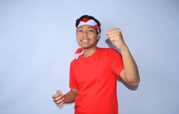 幸せなインドネシアの若い成人は 赤いTシャツとインドネシアの国旗ヘッドバンドを身に着けている平和的な顔で彼の勝利のための勝利をし 彼の拳をラスタリング — ストック写真