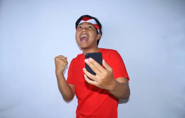 興奮したインドネシアのフリーランサーの男は叫び 右手を上げてイエスのジェスチャーを示し 左手はインドネシア独立記念日に赤いTシャツとヘッドバンドを身に着けている彼の功績を祝う暴徒の携帯電話を保持します — ストック写真