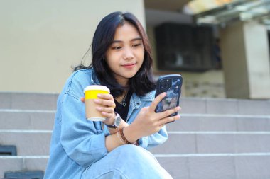 Mutlu Asyalı genç bir kadın akıllı telefondan başvuru kullanıyor, video izliyor, sosyal medya ağında sörf yapıyor ve alışveriş merkezinin merdivenlerinde otururken bir fincan kahve içiyor.