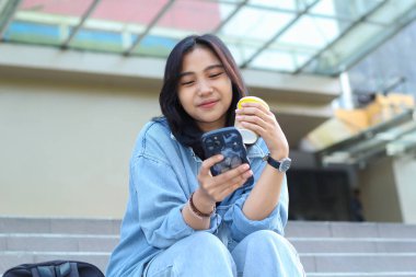 Mutlu Asyalı genç bir kadın akıllı telefondan başvuru kullanıyor, video izliyor, sosyal medya ağında sörf yapıyor ve alışveriş merkezinin merdivenlerinde otururken bir fincan kahve içiyor.