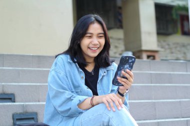 Neşeli Asyalı uzaktan kumandalı kadın gülerek sosyal medyada akıllı telefon sörfü, mobil uygulama, görüntülü sohbet, şehir merdivenlerinde otururken internet indirimi yapmaktan keyif alıyor.