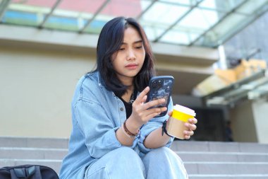 Düşünceli Asyalı genç kadın dijital göçebe akıllı telefonu ciddi ciddi kullanıyor ve bir fincan kahve içiyor sosyal medyada geziniyor ve görüntülü arama yapıyor.