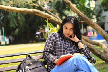 Mutlu ve başarılı Asyalı kız öğrenci parkta kitap okuyarak eğleniyor.