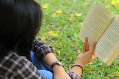 Mutlu ve başarılı Asyalı kız öğrenci parkta kitap okuyarak eğleniyor.