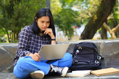 Yorgun ve stresli Asyalı kız öğrenci laptopla çalışıyor ve açık havada ders kitaplarıyla çalışıyor.
