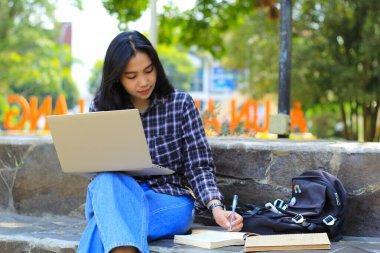 Genç gülümseyen Asyalı kadın dizüstü bilgisayar kullanıyor ve not defteri yazıyor. Neşeli Asyalı öğrenci Webinar izliyor ve dersini çalışıyor.