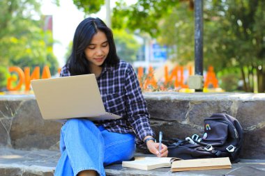 Genç gülümseyen Asyalı kadın dizüstü bilgisayar kullanıyor ve not defteri yazıyor. Neşeli Asyalı öğrenci Webinar izliyor ve dersini çalışıyor.