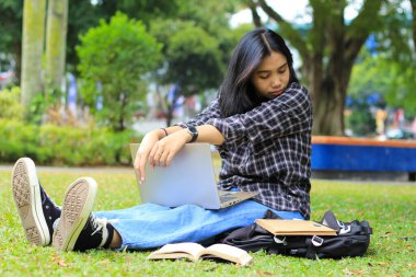 Bilgisayarla uzaktan çalışmak, kitap okumak ve deftere yazmak için internette sörf yapan Asyalı genç bir kadının portresi. Çekici Asyalı öğrenci açık havada okuyor.