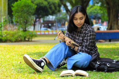 Güzel Asyalı genç kız üniversite öğrencisi şehir parkında defter yazmaya ve kitap okumaya odaklanmış.