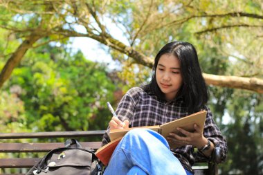 Gülümseyen Asyalı güzel genç kadın şehir parkında deftere liste ve fikir yazmaktan zevk alıyor.