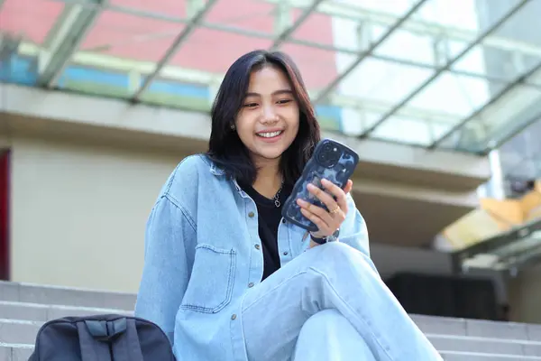 Akıllı telefon uygulaması, görüntülü sohbet, online alışveriş merkezi, sosyal medya ve açık hava merdivenlerinde oturan kaygısız gülümseyen Asyalı genç kadın günlük giyim kuşamı