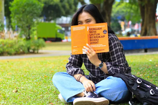 happy young asian woman college student cover up her face with a book looking to camera