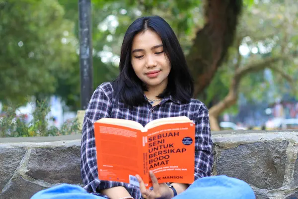 Mutlu, düşünceli Asyalı bir üniversite öğrencisi parkta kitap okuyor, eğitim konsepti.
