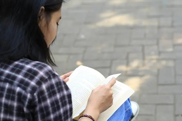 Mutlu ve başarılı Asyalı kız öğrenci parkta kitap okuyarak eğleniyor.