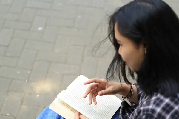 Mutlu ve başarılı Asyalı kız öğrenci parkta kitap okuyarak eğleniyor.