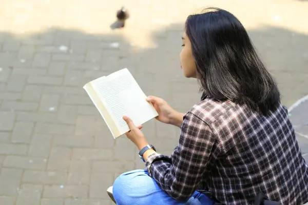 Mutlu ve başarılı Asyalı kız öğrenci parkta kitap okuyarak eğleniyor.