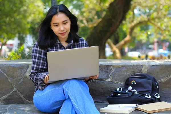 Genç gülümseyen Asyalı kadın dizüstü bilgisayar kullanıyor ve not defteri yazıyor. Neşeli Asyalı öğrenci Webinar izliyor ve dersini çalışıyor.