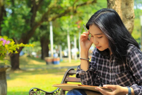 confusing asian young woman thinking an idea and writing in notebook with a pen in nature outdoors area