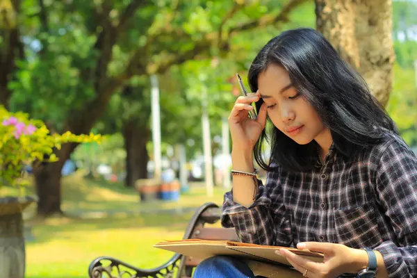 confusing asian young woman thinking an idea and writing in notebook with a pen in nature outdoors area
