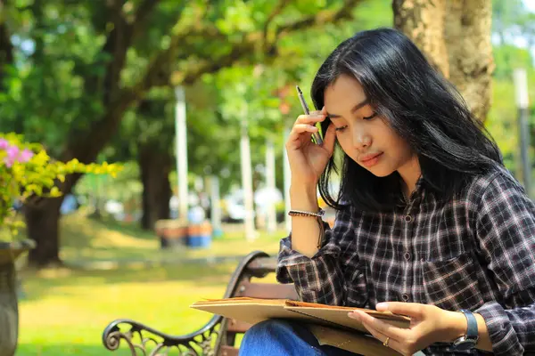 confusing asian young woman thinking an idea and writing in notebook with a pen in nature outdoors area