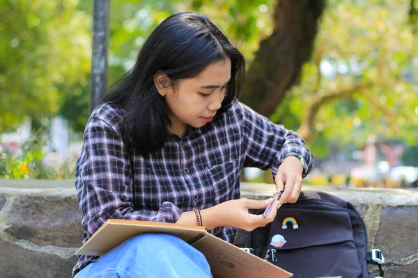 beautiful asian young woman college student focused writing on notebook and reading book in outdoors city park