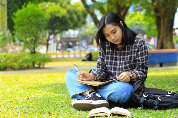 beautiful asian young woman college student focused writing on notebook and reading book in outdoors city park