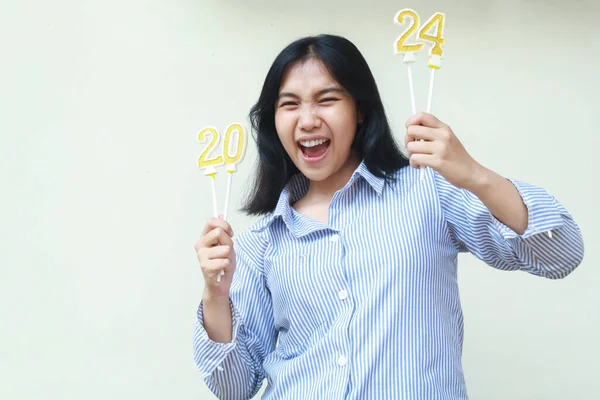 excited asian young woman laugh and scream celebrating new years eve by holding golden candles wearing blue stripes shirt looking at camera isolated on white background