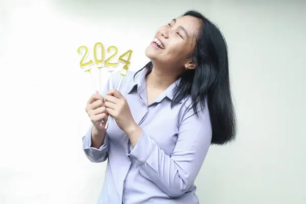 excited asian young business woman wearing grey formal suit looking up with laugh while raising arm holding 2024 figure candle, isolated, female coworker enjoy new years eve celebration