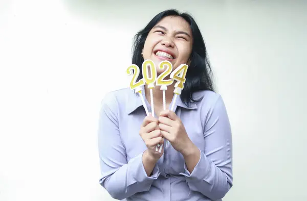Neşeli Asyalı genç iş kadını dişlek bir gülümseme ile gülüyor ve yana bakıyor, 2024 figürlü mumları kaldırıyor, el ele tutuşuyor, münzevi, kadın çalışan gri takım elbise giyiyor yeni yıl arifesini kutluyor.