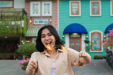 Neşeli genç Asyalı kadın elinde maytap tutarak yılbaşı arifesini bahçe partisiyle renklendirilmiş evin bahçesinde dikiliyor.
