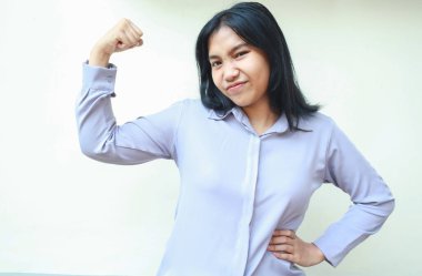 Kaygısız güçlü Asyalı genç iş kadını kollarını kaldırırken kollarını kaldırırken elleri belinde, resmi tişörtü giymiş beyaz arka planda kameraya bakıyor.