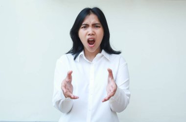 Öfkeli patron Asyalı genç kadın kameraya bağırıyor. Elinde hayali bir ürün tutmaktan memnun değil. Beyaz resmi tişört giymiş, izole edilmiş arka planda dikiliyor.