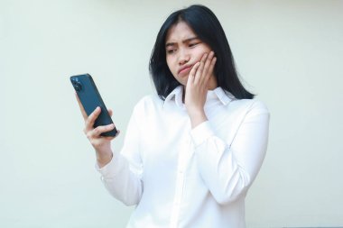 Mutlu olmayan Asyalı bir kadının internet üzerinden sosyal medyada cep telefonuyla dolaşması, online alışveriş hayal kırıklıkları, izole edilmiş arka planda asılı duran resmi beyaz gömlek giymesi.