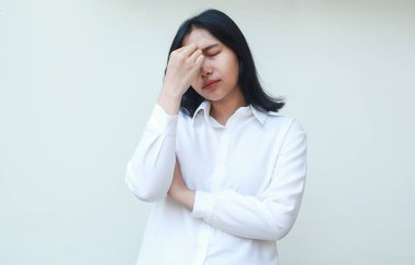 Hüsrana uğramış Asyalı genç iş kadını yöneticisinin görüntüsü. Alnına dokunarak strese giriyor. Beyaz takım elbise giyiyor, kolları bağlı ve gözleri kapalı yorgun duruyor.
