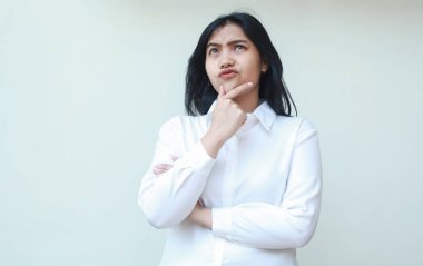 Düşünceli Asyalı genç iş kadınının portresi elleri çenesinde ve kolları bağlı şüpheli bir şekilde yukarı bakıyor, beyaz resmi gömlek giyiyor, ayakta duruyor.