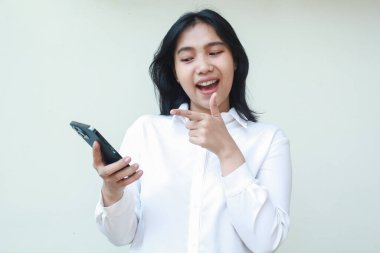 Çekici Asyalı genç satıcı işaret parmağıyla işaret ediyor ve akıllı telefona baş parmak kaldırıyor. Üzerinde beyaz resmi kıyafet tişörtü var. İnternetten alışveriş yapmaktan, sosyal medyayı kullanmaktan, video araması yapmaktan memnun.