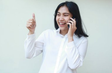 Gülen Asyalı genç girişimcinin beyaz gömlek giyerek, iyi hizmet jesti, tek başına ayakta dikilirken akıllı telefonla konuşurken başını kaldırdığı görüntüsü.