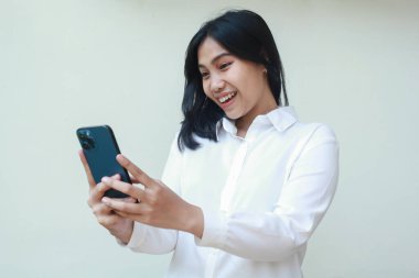 Huzurlu, heyecanlı, beyaz resmi kıyafet giyen Asyalı genç satıcı sosyal medya, online alışveriş, iyi haberler okumak, tecrit edilmiş, kucaklayıcı teknoloji için akıllı telefonu kullanmanın keyfini çıkarın.