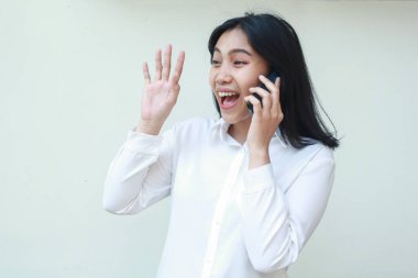 Avuç içi kaldıran Asyalı iş kadını beyaz takım elbise içinde cep telefonuyla konuşurken selam veriyor ve yüzünü çeviriyor. İzole edilmiş arka planda duran hoş geldin hareketi yapıyor.
