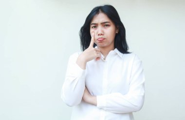 Asyalı mutsuz iş kadınının yanağına dokunduğu görüntüsü. Beyaz tişörtlü, resmi takım elbiseli, kollarını kavuşturmuş kameraya bakıyor, fazla mesai yapmaktan yorulmuş, tek başına ayakta duruyor.