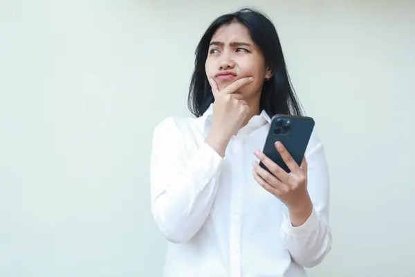Meraklı Asyalı genç bir kadın kafasını çevirmiş cep telefonu uygulamasını Chind 'in üzerinde kullanmayı, internette sörf yapmayı, online alışveriş yapmayı, beyaz arka planda duran beyaz takım elbise giymeyi düşünüyor.