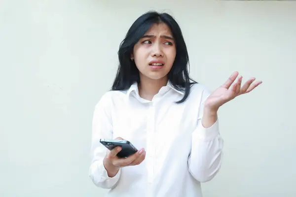 Düşünceli Asyalı kadın yöneticinin portresi boş alana bakıyor kafası karışık, hareket gösteren avuçlarını kaldırıyor, elinde akıllı telefon, beyaz resmi gömlek giyiyor, ayakta duruyor.