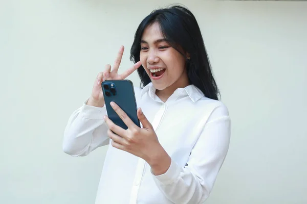 confident young asian woman holding smartphone excited taking selfie of carefree smile face expression with two fingers peace gesture wearing white suit shirt, standing isolated, wireless lifestyle